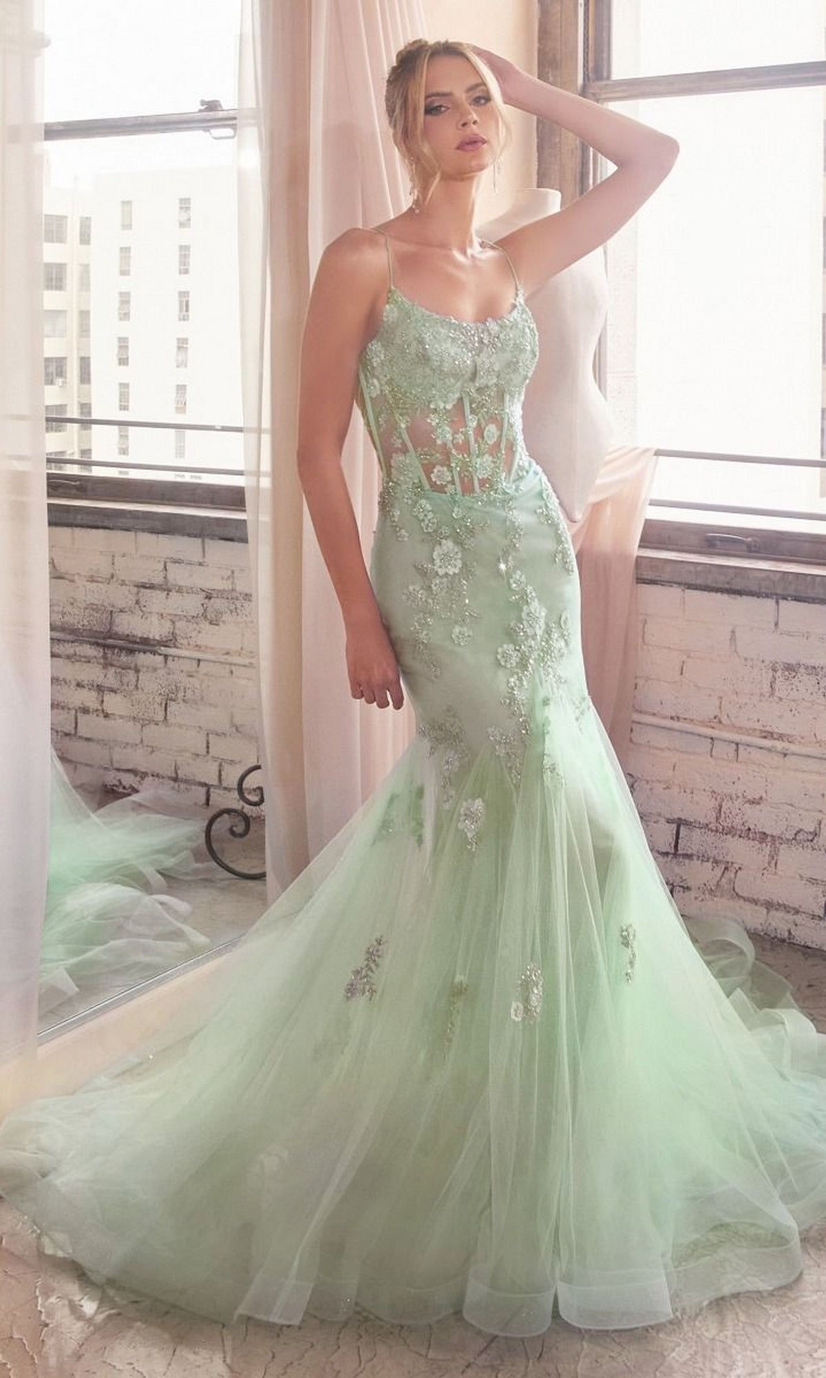 A woman stands by a window wearing the Long Pastel Floral Mermaid Prom Dress D145, a light green, floor-length gown featuring tulle, elegant floral appliqués, sheer panels, and delicate detailing.